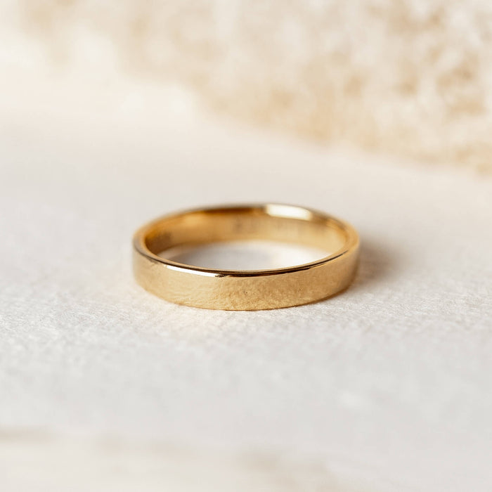 Wide Ring - Paula shown as a simple gold wide band ring on a soft neutral surface with blurred background