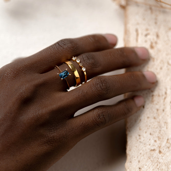 Close-up of a hand wearing the gold Wide Ring - Paula stacked with delicate gemstone and eternity rings on a neutral background