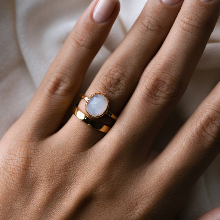 Close-up of a hand wearing the Wide Ring - Paula gold wide band ring styled with a soft neutral fabric background
